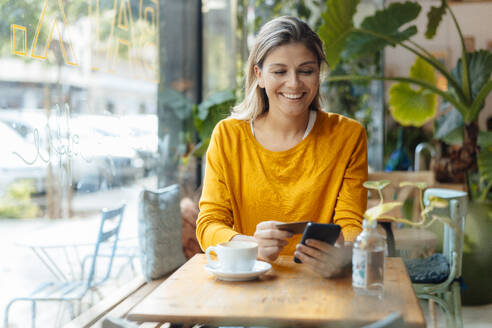 Glückliche Frau mit Kreditkarte und Smartphone in einem Café - JOSEF18308