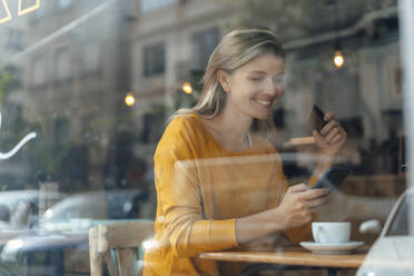 Glückliche Frau mit Kreditkarte und Smartphone in einem Café sitzend - JOSEF18314