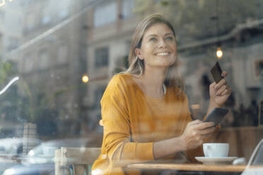 Thoughtful happy woman with credit card and smart phone sitting in cafe - JOSEF18315