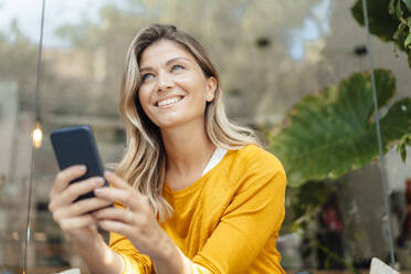 Glückliche Frau mit blondem Haar, die ein Smartphone vor einer Glaswand hält - JOSEF18316