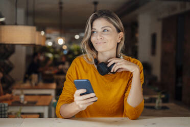 Nachdenklich lächelnde Frau mit Kopfhörern und Mobiltelefon in einem Café sitzend - JOSEF18324