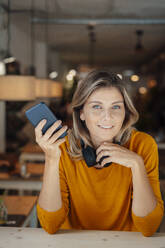 Lächelnde Frau mit blondem Haar hält Smartphone in einem Café - JOSEF18327