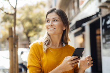 Thoughtful smiling woman with mobile phone outside building - JOSEF18334