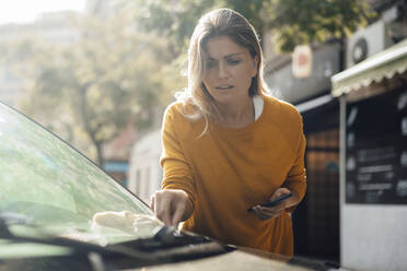 Worried woman picking dirty cloth from windshield of car - JOSEF18335