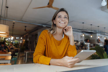 Happy woman holding tablet PC sitting with hand on chin in cafe - JOSEF18337