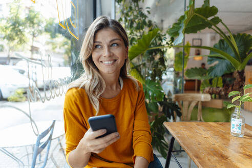 Glückliche Frau mit Smartphone schaut durch ein Fenster in einem Café - JOSEF18339