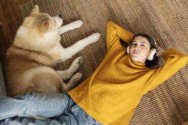 Young woman wearing wireless headphones listening to music and lying on carpet by dog - JCCMF10130