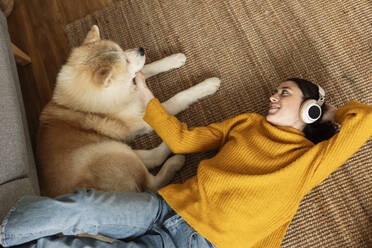 Smiling woman wearing wireless headphones listening to music and stroking dog on carpet at home - JCCMF10131
