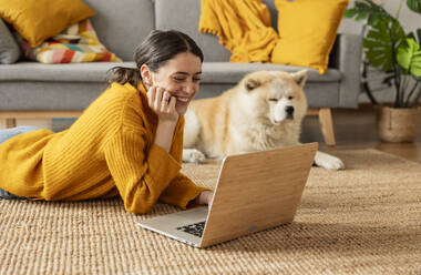 Lächelnde Frau, die auf dem Teppich liegt und einen Laptop mit einem Hund zu Hause benutzt - JCCMF10144