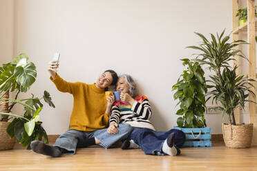 Happy woman taking selfie and toasting coffee cups at home - JCCMF10147