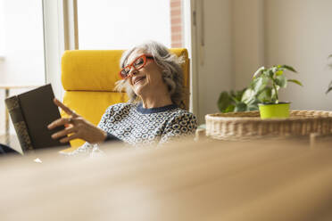 Smiling mature woman reading book sitting in armchair at home - JCCMF10157