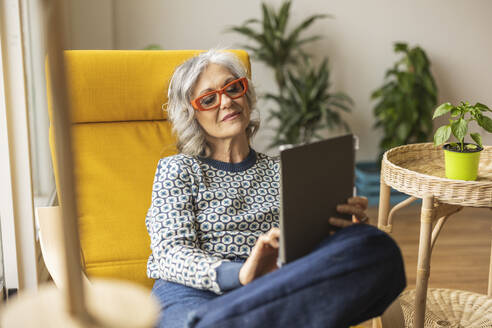 Lächelnde reife Frau, die einen Tablet-PC benutzt und auf einem Stuhl zu Hause sitzt - JCCMF10158