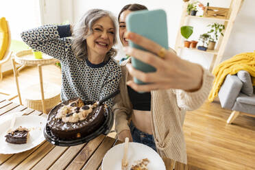 Happy mature woman holding cake with daughter taking selfie through smart phone at home - JCCMF10179