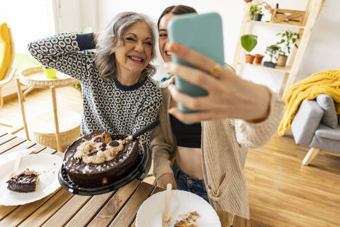 Eine fröhliche Frau mittleren Alters posiert mit ihrer Tochter, während sie ein Selfie mit einem Smartphone macht und zu Hause einen leckeren Kuchen in der Hand hält - JCCMF10179