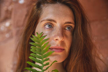 Thoughtful young woman holding fern over face - PCLF00384