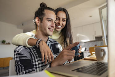 Happy young man discussing with girlfriend over laptop at home office - JCCMF10222