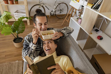 Lächelnder Mann isst einen Snack mit seiner Freundin und liest ein Buch auf dem Sofa zu Hause - JCCMF10227