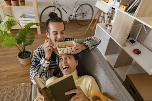 Smiling man eating snack with girlfriend reading book lying on sofa at home - JCCMF10227