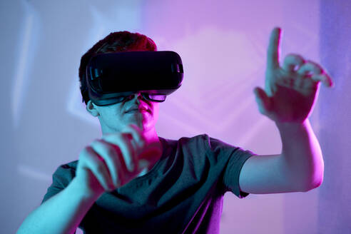 Junger Mann mit Virtual-Reality-Simulator gestikuliert vor einer Neonwand - ABIF01911