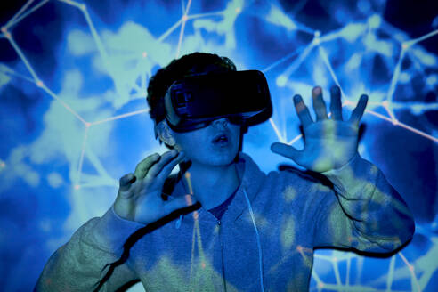 Young man wearing virtual reality simulator gesturing in front of illuminated wall - ABIF01945