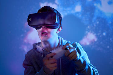 Junger Mann mit VR-Brille hält Spiel-Controller mit Projektionsfläche im Hintergrund - ABIF01951