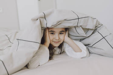 Lächelndes süßes Mädchen liegt unter einer Decke auf dem Bett - VBUF00290