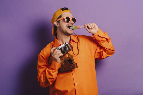 Stilvoller Mann in orangefarbenem Hemd mit Kamera und Eiscreme vor lila Hintergrund - VSNF00663