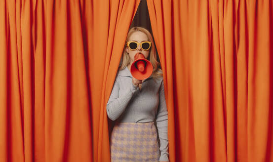 Woman talking through megaphone standing amidst orange curtains - VSNF00666