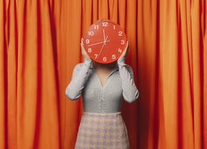 Frau hält rote Uhr über Gesicht vor orangefarbenem Vorhang - VSNF00670