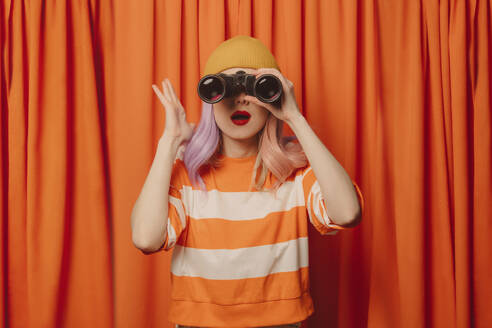 Überraschte Frau, die durch ein Fernglas vor einem orangefarbenen Vorhang schaut - VSNF00676