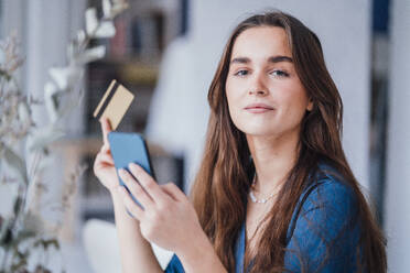 Confident young businesswoman with mobile phone and credit card in office - JOSEF18347