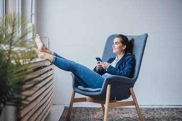 Glückliche junge Geschäftsfrau mit Smartphone in einem Sessel sitzend - JOSEF18394
