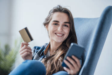 Smiling businesswoman with mobile phone and credit card sitting in chair - JOSEF18399