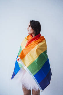 Thoughtful young woman with rainbow flag standing in front of wall - JOSEF18440