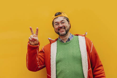 Happy mature man gesturing peace sign against yellow background - VSNF00680