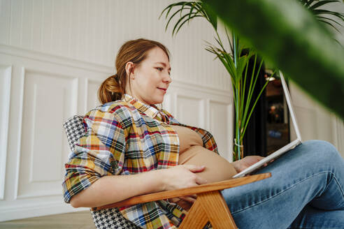 Lächelnde schwangere Frau sitzt mit Laptop auf einem Stuhl zu Hause - MDOF01043