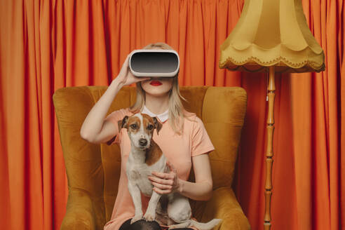 Frau mit VR-Brille sitzt mit Hund vor orangefarbenen Vorhängen - VSNF00682
