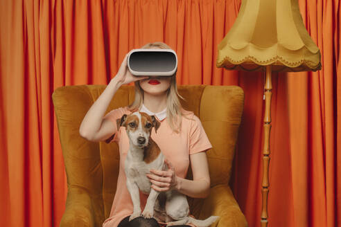 Woman wearing VR glasses sitting with dog in front of orange curtains - VSNF00682