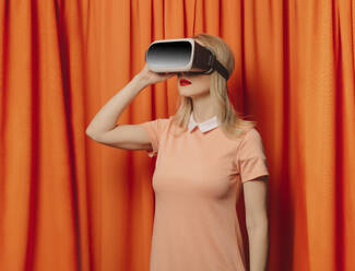 Frau mit Virtual-Reality-Simulator vor orangefarbenen Vorhängen stehend - VSNF00683