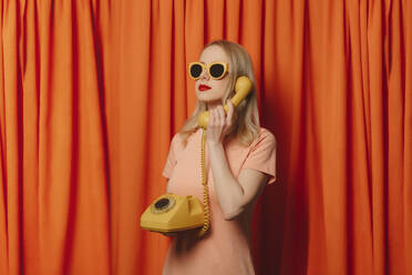 Frau mit Sonnenbrille spricht über Festnetztelefon vor orangefarbenen Vorhängen - VSNF00685