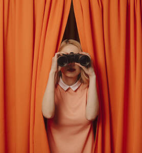 Frau schaut durch ein Fernglas inmitten von orangefarbenen Vorhängen - VSNF00686