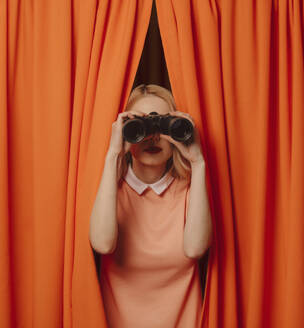 Woman looking through binoculars amidst orange curtains - VSNF00686