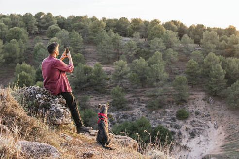 Hund sitzt neben einem Mann, der mit seinem Smartphone bei Sonnenuntergang die Landschaft fotografiert - MGRF00928