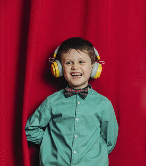 Happy boy wearing wireless headphones standing in front of red curtain - VSNF00692