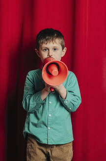 Boy talking in megaphone standing in front of red curtain - VSNF00694