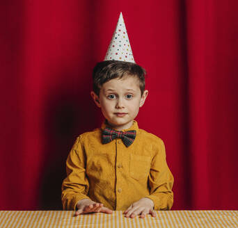 Boy in birthday hat sitting at table in front of red curtain - VSNF00697