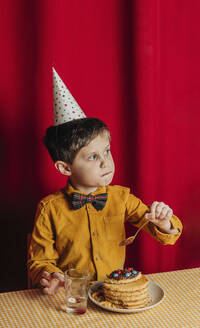 Boy eating birthday cake at table in front of red curtain - VSNF00699