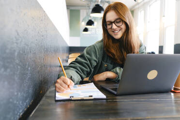 Happy young freelancer working with documents at table in cafe - ASGF03527