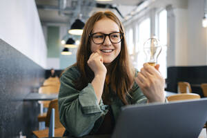 Happy freelancer with light bulb sitting in cafe - ASGF03534