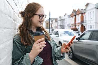 Smiling young woman standing with disposable coffee cup and using smart phone - ASGF03545